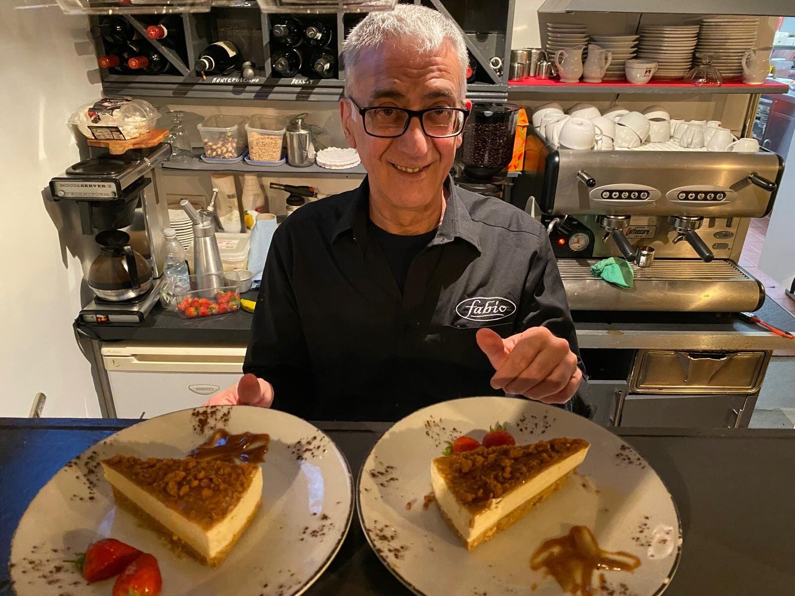Angelo, Fabio's dessert specialist, smiling behind the bar in Swindon with two plated slices of cheesecake and strawberries