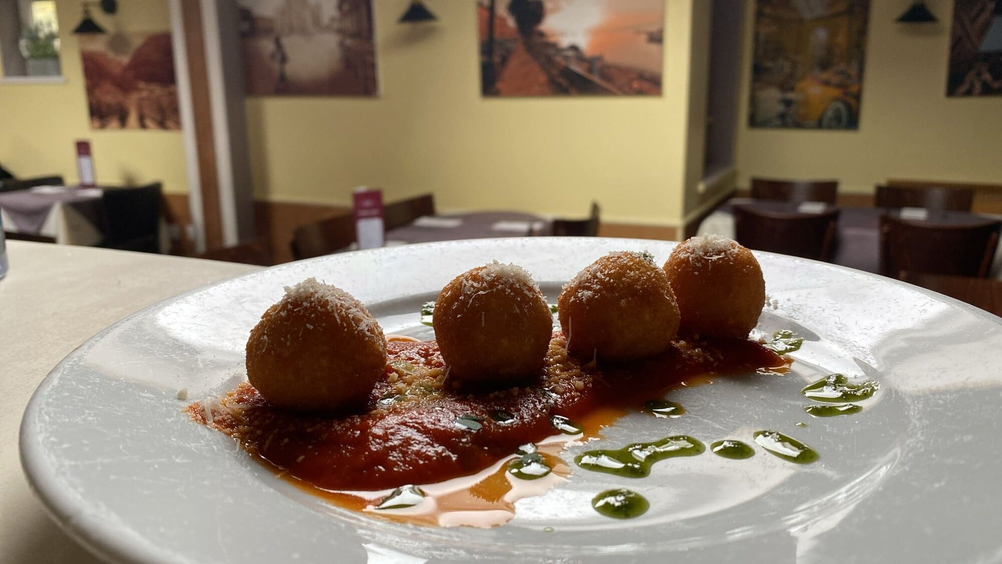 Four golden, crumb-fried balls topped with grated Parmesan on tomato sauce with basil oil, with Fabio's dining room behind